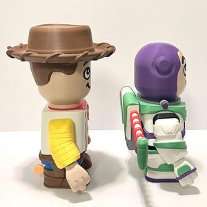 May include: Two character figurines, Woody and Buzz Lightyear, from the animated film Toy Story. Woody wears a brown cowboy hat, a yellow sleeve, and a red neckerchief. Buzz Lightyear has a purple helmet, green and white spacesuit, and red and white jetpack.