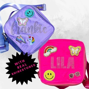 Peut inclure: Deux sacs à déjeuner, l'un violet et l'autre rose, avec les noms personnalisés "Frankie" et "Lila" en strass. Les deux sacs présentent divers patchs colorés, notamment des papillons, des arcs-en-ciel et des visages souriants.