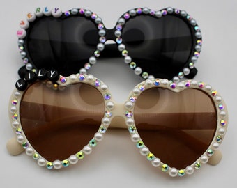 May include: Two pairs of heart-shaped sunglasses with pearl embellishments. The top pair has black lenses and the word "EVERLY" spelled out in colorful letters. The bottom pair has brown lenses and the word "LILLY" spelled out in black letters.