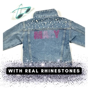 May include: A blue denim jacket with the name "MACY" spelled out in pink and purple rhinestones. The jacket is decorated with white pearl-like beads.