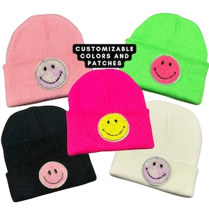 Könnte beinhalten: Fünf Beanies mit verschiedenen bunten Smiley-Patches. Die Mützen sind rosa, grün, pink, schwarz und weiß. Der Text "CUSTOMIZABLE COLORS AND PATCHES" befindet sich auf einem weißen Rechteck in der Mitte des Bildes.