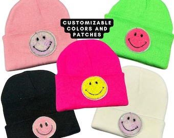 May include: Five beanie hats with different colored smiley face patches. The hats are pink, green, hot pink, black, and white. The text "CUSTOMIZABLE COLORS AND PATCHES" is on a white rectangle in the center of the image.