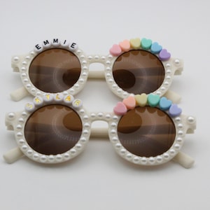 May include: Two pairs of white pearl-embellished sunglasses with brown lenses. The top pair has the name "EMMIE" spelled out in black letters on the frame. The bottom pair has the name "SKYLAR" spelled out in gold letters on the frame. Both pairs have colorful heart-shaped beads on the frames.