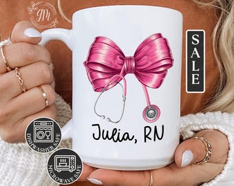 May include: White ceramic mug with a pink bow and stethoscope design. The mug says "Julia, RN" and has dishwasher and microwave safe labels.