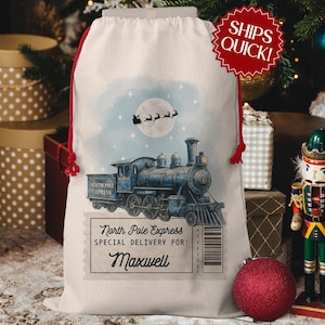May include: A cream-colored Christmas gift bag with a watercolor illustration of a blue steam train labeled "North Pole Express." The bag features a red drawstring and the text "Special Delivery for: Maxwell."