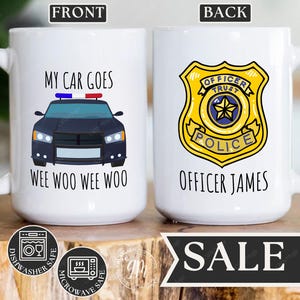 May include: White ceramic mug with a black and blue police car graphic and the text "My car goes wee woo wee woo". The back of the mug has a yellow police badge with the text "Officer Trust Police" and "Officer James".