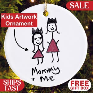 May include: White ceramic ornament with a gold string for hanging. The ornament features a child's drawing of two figures with crowns and dresses, with the text "Mommy + Me". The words "Kids Artwork Ornament" are also visible.