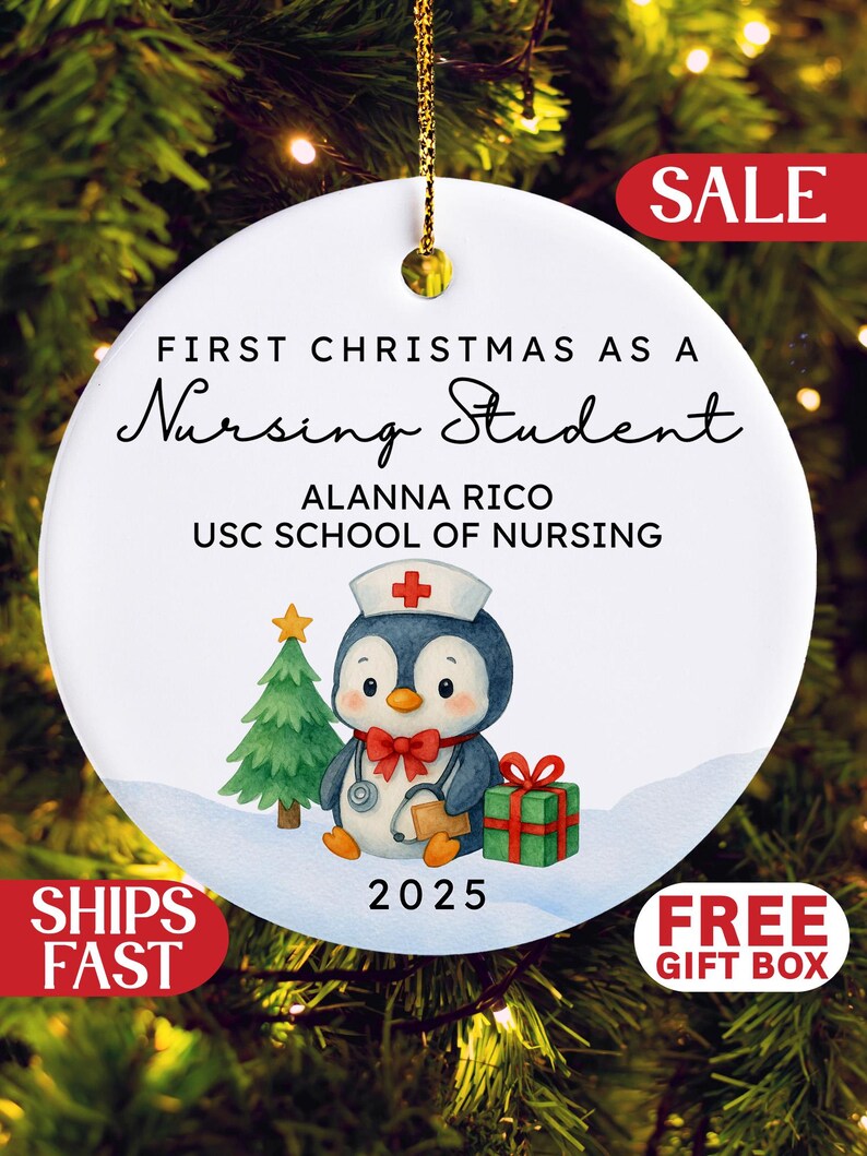 May include: White ceramic Christmas ornament with gold hanging string. The ornament features a penguin in a nurse's hat, a Christmas tree, and a gift. Text reads "First Christmas as a Nursing Student" and "2025".