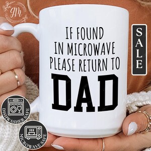 May include: White ceramic mug with black text that reads "If found in microwave please return to Dad". The mug is being held by a person's hands.