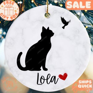 May include: A round, white ceramic ornament with a black cat silhouette and a bird holding a sprig. The ornament has the name "Lola" with a red heart. The word "SALE" is in an orange banner. A gift box icon is visible.