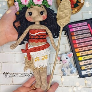 May include: Crocheted amigurumi doll of Moana from the Disney movie Moana. The doll is wearing a brown and orange dress with a white crocheted skirt and a flower crown. The doll is holding a brown crocheted spear. There is a small crocheted pig beside the doll.