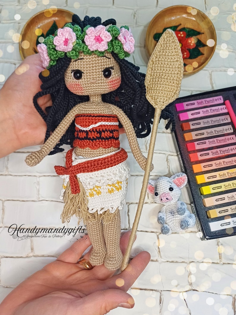 May include: Crocheted amigurumi doll of Moana from the Disney movie Moana. The doll is wearing a brown and orange dress with a white crocheted skirt and a flower crown. The doll is holding a brown crocheted spear. There is a small crocheted pig beside the doll.