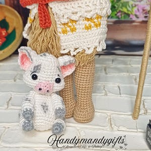 May include: A crocheted doll with a red and white skirt and a small crocheted pig. The doll is standing on a white brick surface. The doll is wearing a red headband and has black hair. The pig is white with black spots and has a pink nose.