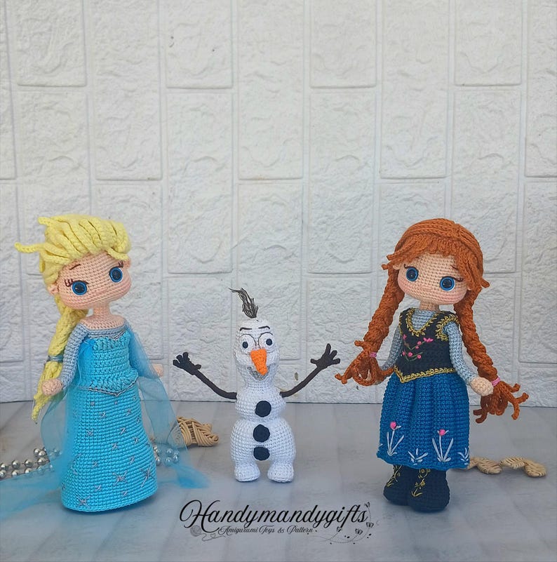May include: Three handmade crochet dolls: Elsa in a blue dress, Anna in a blue dress, and Olaf the snowman. Elsa has blonde hair, Anna has red braids, and Olaf is white with a carrot nose. The dolls are on a light surface.