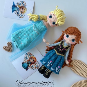May include: Two handmade crochet dolls, one with blonde hair and a blue dress, the other with red hair and a blue dress. The dolls are lying on a white surface with two heart-shaped cards and the text "Handymandy gifts".