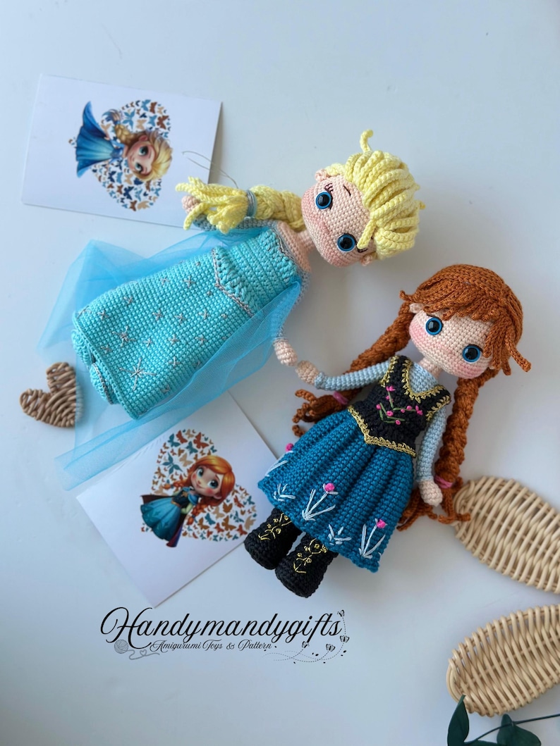 May include: Two handmade crochet dolls, one with blonde hair and a blue dress, the other with red hair and a blue dress. The dolls are lying on a white surface with two heart-shaped cards and the text "Handymandy gifts".