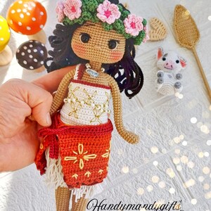 Peut inclure: Poup&eacute;e en crochet aux cheveux bruns, portant une couronne de fleurs roses, un haut blanc avec des broderies dor&eacute;es, une jupe rouge avec des broderies jaunes et des sandales blanches. La poup&eacute;e tient un petit cochon en crochet.