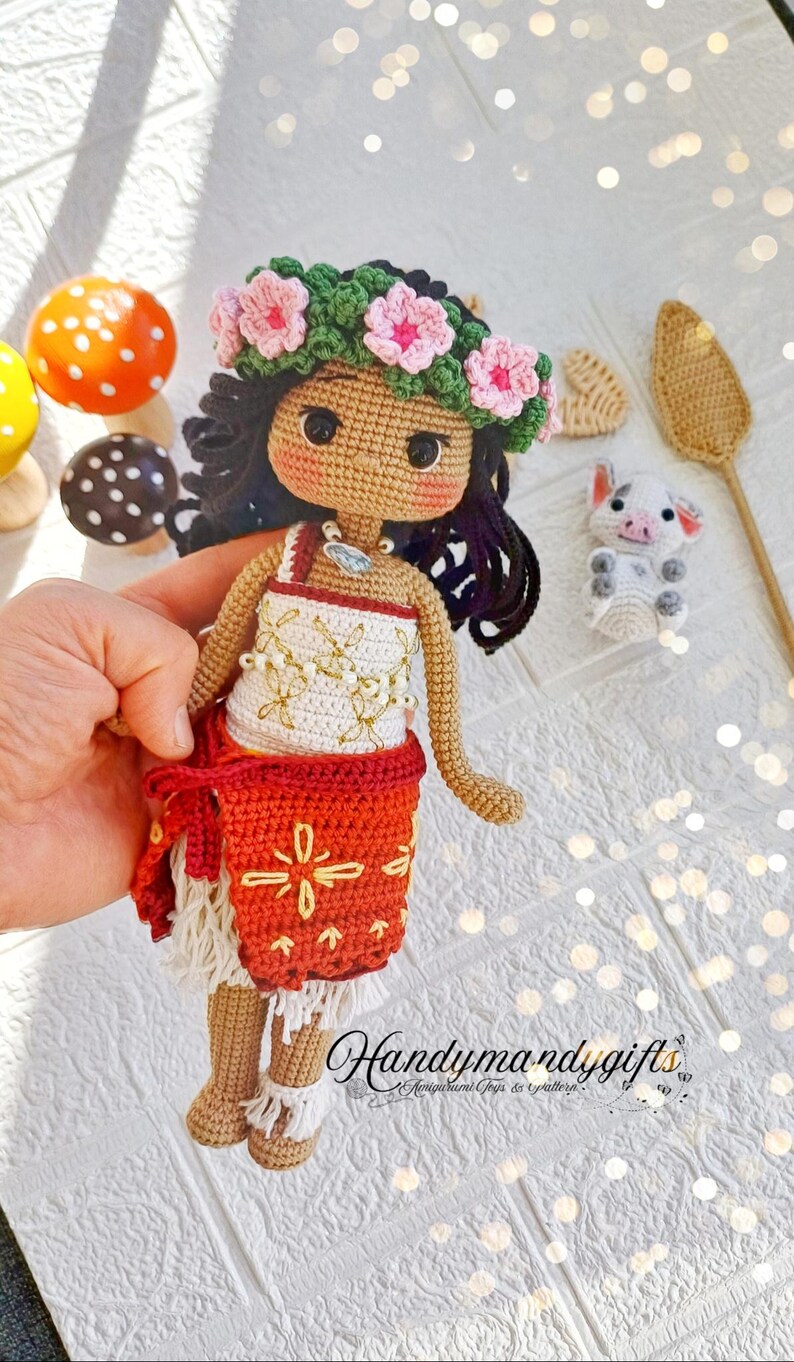 Peut inclure: Poup&eacute;e en crochet aux cheveux bruns, portant une couronne de fleurs roses, un haut blanc avec des broderies dor&eacute;es, une jupe rouge avec des broderies jaunes et des sandales blanches. La poup&eacute;e tient un petit cochon en crochet.