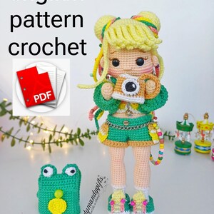 Puede incluir: Muñeca de ganchillo hecha a mano con cabello rubio, vestida con un atuendo verde y sosteniendo una cámara. La muñeca tiene un accesorio en forma de rana y el texto "english pattern crochet" es visible. La muñeca mide unos 15 cm de alto.