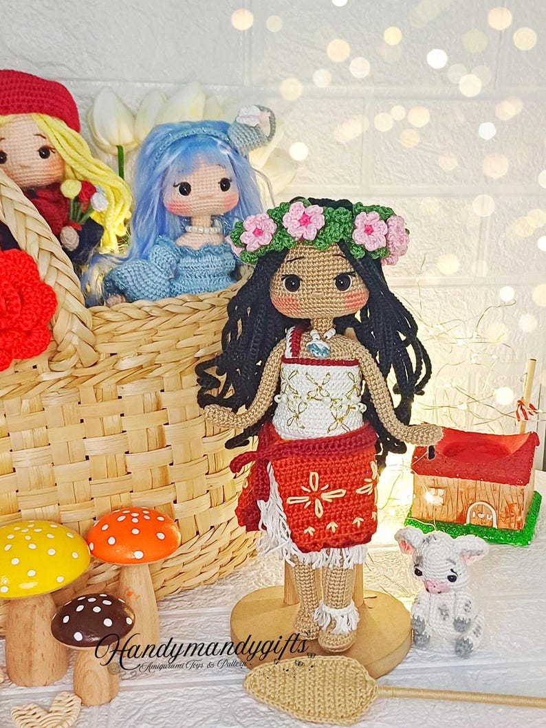 Peut inclure: Poup&eacute;e en crochet avec des cheveux bruns, une couronne de fleurs et une robe rouge et blanche. La poup&eacute;e est debout sur une base en bois et tient un petit agneau en crochet. La poup&eacute;e est entour&eacute;e d'autres poup&eacute;es et jouets en crochet, dont un champignon et un panier.