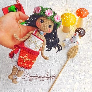 Peut inclure: Poup&eacute;e amigurumi en crochet avec des cheveux bruns, une couronne de fleurs et une jupe rouge et blanche. La poup&eacute;e tient un b&acirc;ton brun avec une extr&eacute;mit&eacute; en crochet. La poup&eacute;e est entour&eacute;e de petits champignons en crochet.