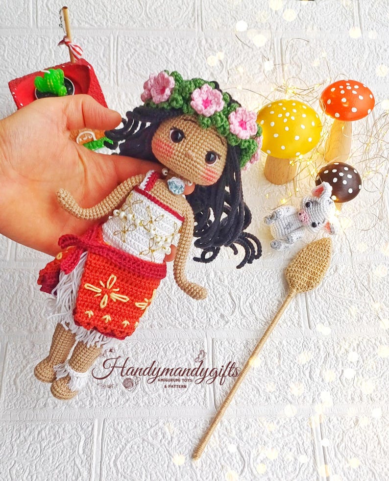 Peut inclure: Poup&eacute;e amigurumi en crochet avec des cheveux bruns, une couronne de fleurs et une jupe rouge et blanche. La poup&eacute;e tient un b&acirc;ton brun avec une extr&eacute;mit&eacute; en crochet. La poup&eacute;e est entour&eacute;e de petits champignons en crochet.