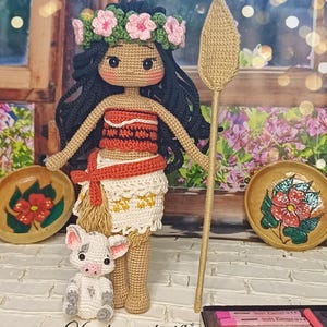 May include: Crocheted amigurumi doll with brown skin, black hair, and a flower crown. The doll is wearing a red and white dress and holding a brown stick. A small crocheted pig is sitting in front of the doll.