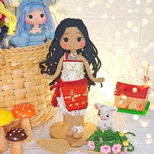 Peut inclure: Poup&eacute;e en crochet aux longs cheveux noirs, v&ecirc;tue d'une robe rouge et blanche, debout sur une base en bois. La poup&eacute;e est entour&eacute;e d'autres objets en crochet, dont un petit agneau blanc, un champignon et une petite maison.