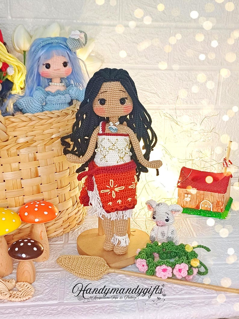 Peut inclure: Poup&eacute;e en crochet aux longs cheveux noirs, v&ecirc;tue d'une robe rouge et blanche, debout sur une base en bois. La poup&eacute;e est entour&eacute;e d'autres objets en crochet, dont un petit agneau blanc, un champignon et une petite maison.
