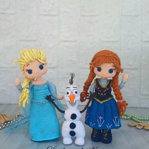May include: Crocheted dolls of Elsa, Anna, and Olaf from the movie Frozen. Elsa wears a blue dress with a cape, Anna has a blue dress with a black bodice, and Olaf is a white snowman with black buttons and stick arms. The dolls are handmade.
