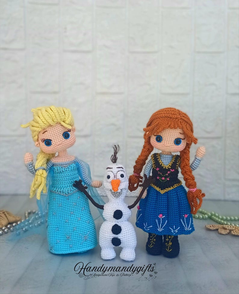 May include: Crocheted dolls of Elsa, Anna, and Olaf from the movie Frozen. Elsa wears a blue dress with a cape, Anna has a blue dress with a black bodice, and Olaf is a white snowman with black buttons and stick arms. The dolls are handmade.