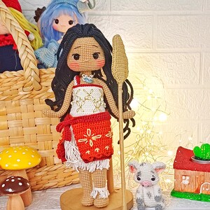 Peut inclure: Poup&eacute;e amigurumi en crochet avec une peau brune, des cheveux noirs et une robe rouge et blanche. La poup&eacute;e tient un b&acirc;ton en bois et se tient sur une base en bois. Un petit cochon en crochet est assis devant la poup&eacute;e.