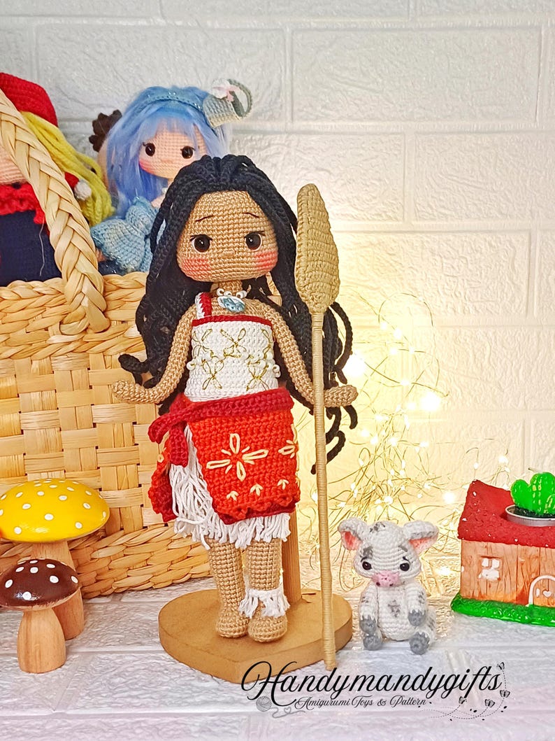 Peut inclure: Poup&eacute;e amigurumi en crochet avec une peau brune, des cheveux noirs et une robe rouge et blanche. La poup&eacute;e tient un b&acirc;ton en bois et se tient sur une base en bois. Un petit cochon en crochet est assis devant la poup&eacute;e.