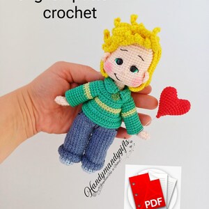 Puede incluir: Muñeco de ganchillo de un niño con pelo amarillo, que lleva un jersey verde y pantalones azules, sosteniendo un corazón rojo. El texto "english pattern crochet" está encima del muñeco. Un icono PDF está en la esquina inferior derecha.