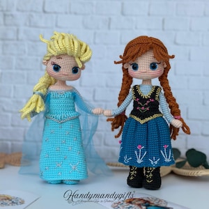 May include: Two handmade crochet dolls, one with blonde hair in a blue dress and the other with red hair in a blue dress. The dolls are holding hands. The dolls are on a white brick background.