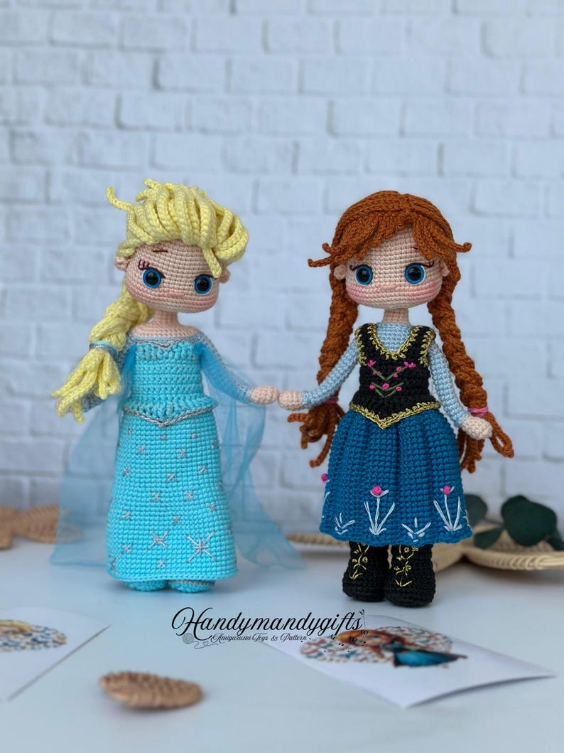 May include: Two handmade crochet dolls, one with blonde hair in a blue dress and the other with red hair in a blue dress. The dolls are holding hands. The dolls are on a white brick background.