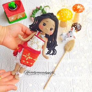 Peut inclure: Poup&eacute;e en crochet avec une peau brune, des cheveux noirs et une robe blanche et rouge. La poup&eacute;e tient un b&acirc;ton en bois avec un sommet en crochet brun. La poup&eacute;e est entour&eacute;e de petits champignons en crochet et d'une petite maison en crochet.