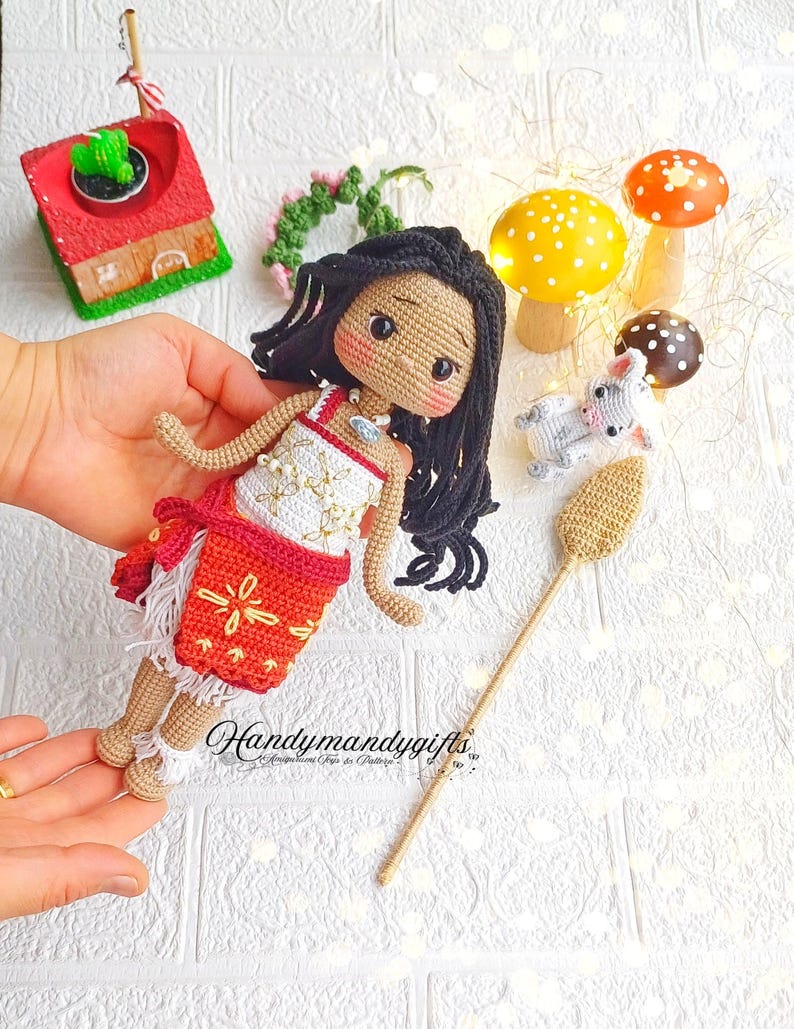 Peut inclure: Poup&eacute;e en crochet avec une peau brune, des cheveux noirs et une robe blanche et rouge. La poup&eacute;e tient un b&acirc;ton en bois avec un sommet en crochet brun. La poup&eacute;e est entour&eacute;e de petits champignons en crochet et d'une petite maison en crochet.