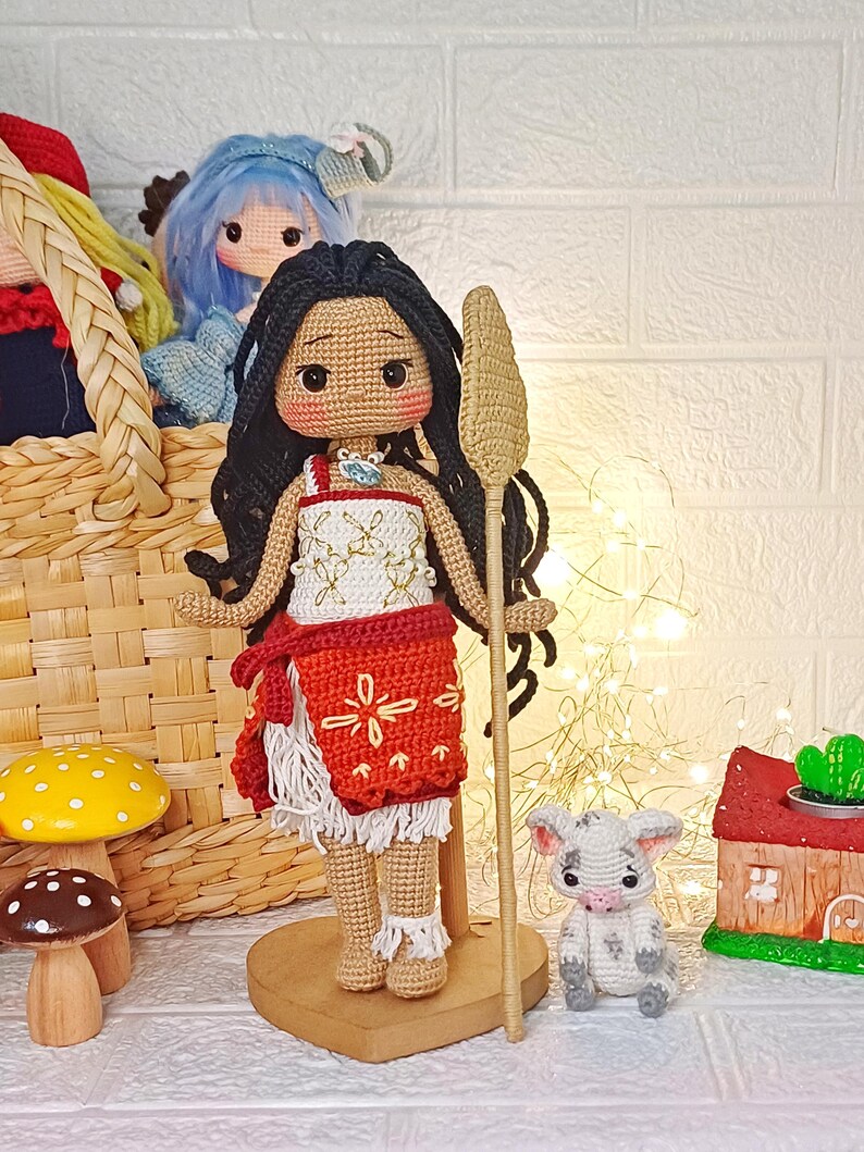Peut inclure: Poup&eacute;e en crochet avec une peau brune, des cheveux noirs et une robe rouge et blanche. La poup&eacute;e tient un b&acirc;ton en bois et se tient sur une base en bois en forme de c&oelig;ur. Un petit cochon en crochet est assis devant la poup&eacute;e.