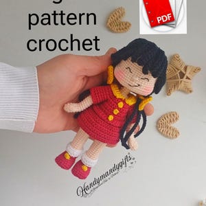 Può includere: Bambola all'uncinetto fatta a mano con un vestito rosso con dettagli gialli, capelli neri e guance rosa. La bambola è tenuta in mano, con le parole "english pattern crochet" sullo sfondo. È visibile anche un'icona PDF.