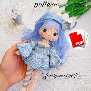Puede incluir: Patrón de muñeca de crochet en formato PDF. La muñeca lleva un vestido azul y tiene el pelo azul. La muñeca sostiene una pequeña regadera. El patrón está disponible para comprar en Etsy.
