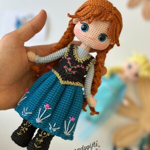 May include: Crocheted doll featuring a character with red braided hair, blue eyes, and a blue dress with floral embroidery. The doll has black boots with gold accents. The doll is held in a hand. The text "Handymandygifts" is visible.