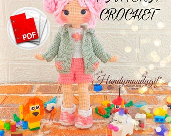 May include: Crochet doll pattern in PDF format. The doll is wearing a green jacket, white shirt, pink shorts, and pink shoes. The doll has pink hair and brown eyes. The doll is standing on a brick surface with colorful toy blocks.