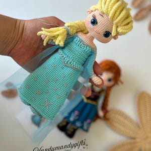 May include: A handmade crochet doll of a character with blonde hair and a blue dress. The doll has large blue eyes and is held in a hand. The dress has a star pattern. The doll is part of a set of two dolls. The text "Handymandygifts" is visible.