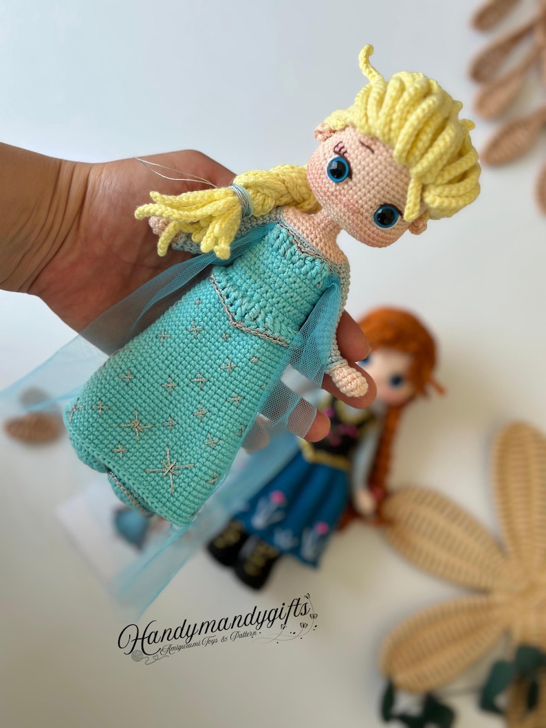 May include: A handmade crochet doll of a character with blonde hair and a blue dress. The doll has large blue eyes and is held in a hand. The dress has a star pattern. The doll is part of a set of two dolls. The text "Handymandygifts" is visible.