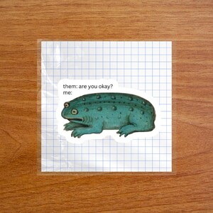 May include: A sticker featuring a teal, textured toad illustration with the printed text "them: are you okay? me:" positioned above the creature. The sticker is placed on a square sheet of white paper with a blue grid pattern.