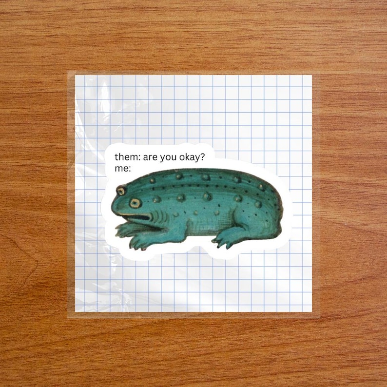 May include: A sticker featuring a teal, textured toad illustration with the printed text "them: are you okay? me:" positioned above the creature. The sticker is placed on a square sheet of white paper with a blue grid pattern.