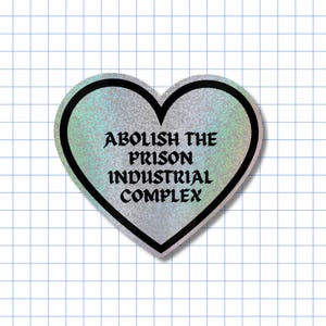 May include: A heart-shaped sticker with a holographic silver finish and a thick black border. The text "ABOLISH THE PRISON INDUSTRIAL COMPLEX" is printed in the centre in a bold, black, gothic-style font against the shimmering background.