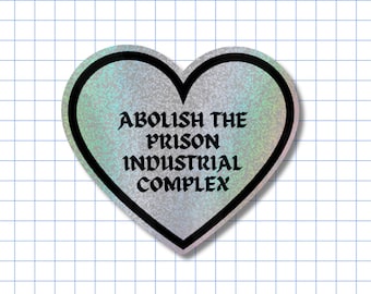 May include: A heart-shaped sticker with a holographic silver finish and a thick black border. The text "ABOLISH THE PRISON INDUSTRIAL COMPLEX" is printed in the center in a bold, black, gothic-style font against the shimmering background.