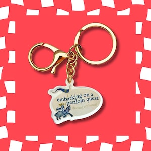 May include: A gold-toned metal keyring featuring a lobster clasp and a split ring attached to an oval-shaped acrylic charm. The charm displays a graphic of a person riding a horse with a flag and the text "embarking on a perilous quest (leaving my house)".